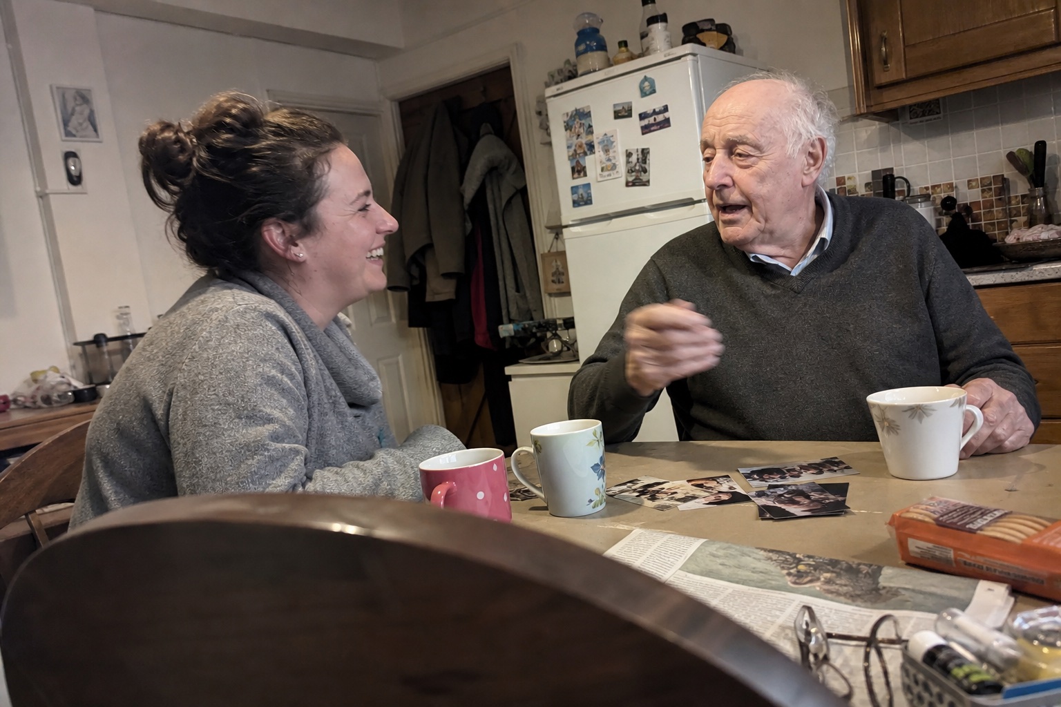 A granddaughter laughing with her grandfather while looking through family photographs at his kitchen table.
