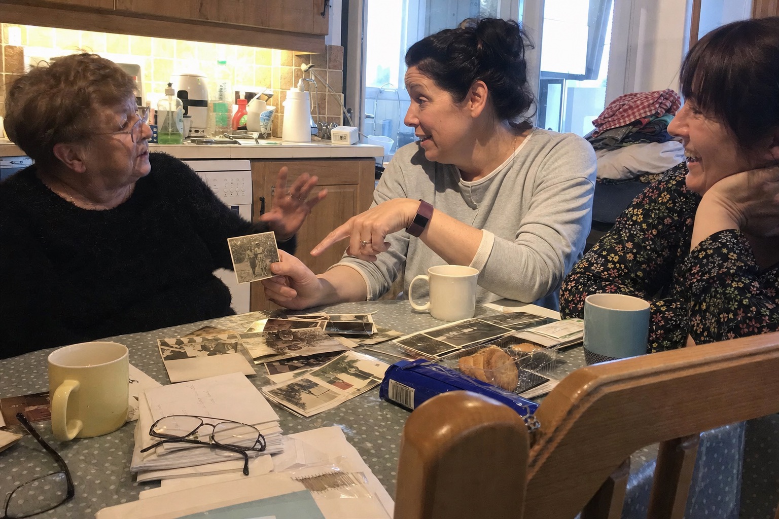 An older woman and two relatives comparing stories while looking through old family photographs at a kitchen table.